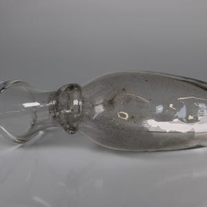 May include: A clear glass bottle with a bulbous body and a long, narrow neck. The bottle has a stopper and is lying horizontally. The glass has a slightly smoky appearance, and the bottle is set against a neutral background.