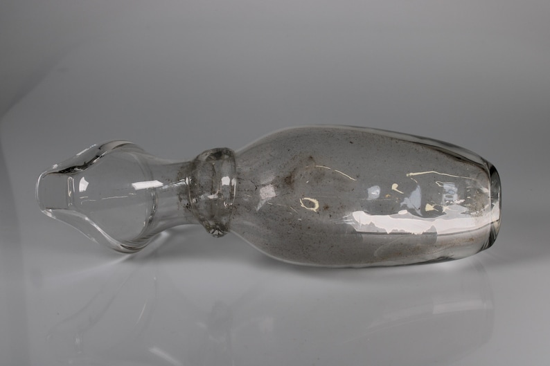 May include: A clear glass bottle with a bulbous body and a long, narrow neck. The bottle has a stopper and is lying horizontally. The glass has a slightly smoky appearance, and the bottle is set against a neutral background.