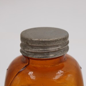 May include: Close-up of a vintage amber glass bottle with a textured, silver-colored metal cap. The bottle has a rounded shape and a visible crack. The cap is tightly sealed, suggesting the bottle's contents are preserved.