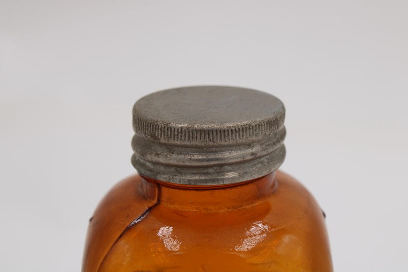 May include: Close-up of a vintage amber glass bottle with a textured, silver-colored metal cap. The bottle has a rounded shape and a visible crack. The cap is tightly sealed, suggesting the bottle's contents are preserved.
