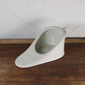 Könnte beinhalten: Ein weißes Porzellan-Nachttopf mit Griff. Der Nachttopf hat eine gebogene Form mit einer breiten Öffnung und einem schrägen Boden. Er steht auf einer Holzoberfläche.