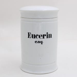 Op de afbeelding: Een witte keramische pot met deksel, met het woord "Eucerin" in zwarte letters. De pot heeft een cilindrische vorm met een licht uitlopende basis en een afgeronde bovenkant.