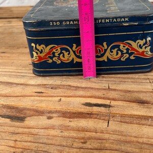May include: A vintage rectangular tin, navy blue with gold and red decorative accents. The tin reads "250 GRAM" and "IFENTABAK". The tin is approximately 4 inches tall, and is on a wooden surface.