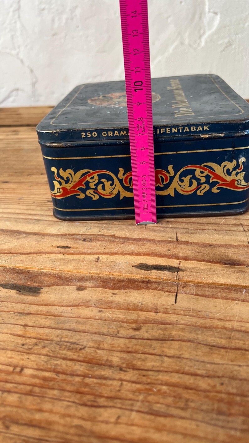May include: A vintage rectangular tin, navy blue with gold and red decorative accents. The tin reads "250 GRAM" and "IFENTABAK". The tin is approximately 4 inches tall, and is on a wooden surface.