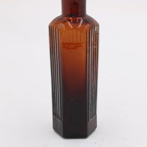 Puede incluir: Una botella de vidrio marr&oacute;n vintage con forma rectangular y detalles acanalados verticales. La botella es de color &aacute;mbar oscuro y tiene una base hexagonal. La botella est&aacute; vac&iacute;a y se alza sobre un fondo blanco.