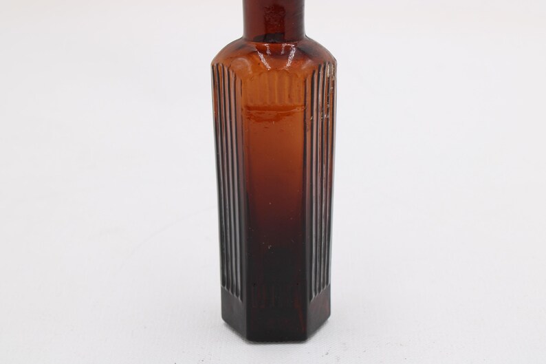 Puede incluir: Una botella de vidrio marr&oacute;n vintage con forma rectangular y detalles acanalados verticales. La botella es de color &aacute;mbar oscuro y tiene una base hexagonal. La botella est&aacute; vac&iacute;a y se alza sobre un fondo blanco.