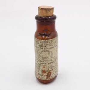 May include: A small, antique brown glass bottle with a cork stopper. The bottle has a label with faded text, including the words "TABLOID" and "Arsenico Comp". The bottle is likely from the late 19th or early 20th century.