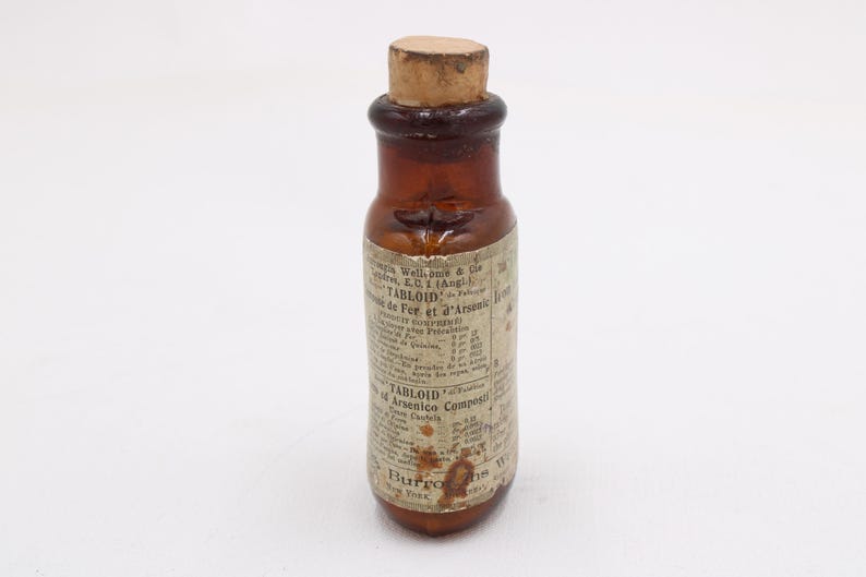 May include: A small, antique brown glass bottle with a cork stopper. The bottle has a label with faded text, including the words "TABLOID" and "Arsenico Comp". The bottle is likely from the late 19th or early 20th century.