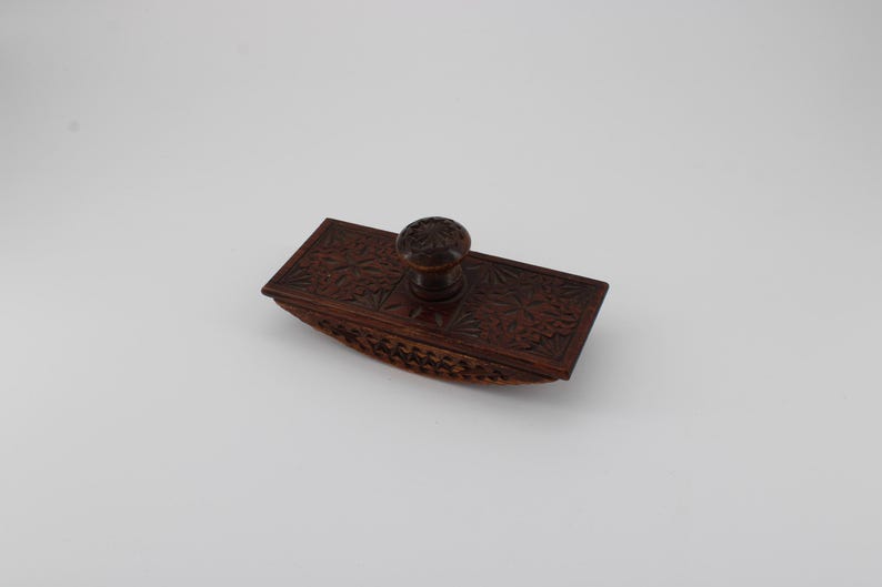 May include: A dark brown, rectangular ink blotter with a carved floral design. The blotter has a rounded base and a round handle on top. The wood has a rich, warm tone, and the carving is detailed and intricate.