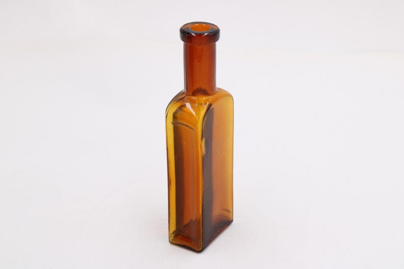 May include: Amber-colored glass bottle with a rectangular body and a narrow neck. The bottle has a vintage aesthetic, with a slightly rounded base and a dark amber hue. The glass appears to be transparent, allowing light to pass through.