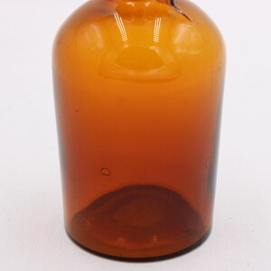 May include: A vintage amber glass bottle with a rounded body and a narrow neck. The bottle has a smooth, glossy surface and a dark brown color that fades to a lighter amber towards the top. The bottle is empty.