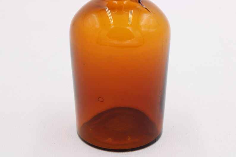 May include: A vintage amber glass bottle with a rounded body and a narrow neck. The bottle has a smooth, glossy surface and a dark brown color that fades to a lighter amber towards the top. The bottle is empty.