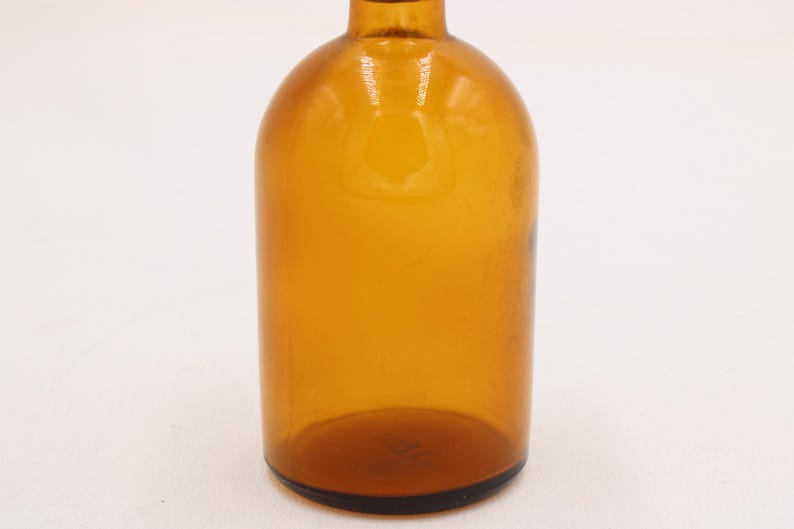 May include: Amber glass bottle with a rounded body and a narrow neck. The bottle has a dark brown base and a smooth, glossy surface. The bottle is empty and appears to be vintage.