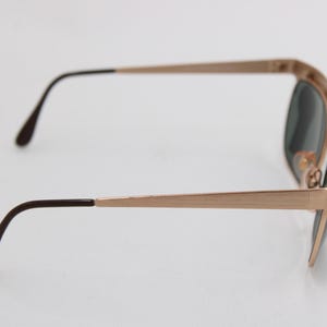 May include: Rose gold-toned sunglasses with dark lenses. The frames have a rectangular shape with a subtle, brushed finish. The temples are slender and end with dark brown tips. A classic accessory.