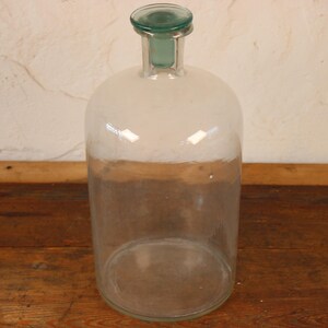 May include: A large, clear glass bottle with a rounded body and a narrow neck. The bottle has a green glass stopper and a white and brown band around the middle. The bottle is sitting on a wooden surface.