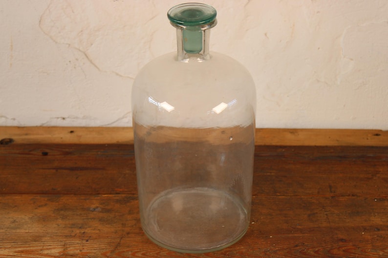 May include: A large, clear glass bottle with a rounded body and a narrow neck. The bottle has a green glass stopper and a white and brown band around the middle. The bottle is sitting on a wooden surface.