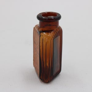 K&ouml;nnte beinhalten: Bernsteinfarbene, rechteckige Glasflasche mit rundem Hals. Die Flasche hat vertikale Rippendetails an den Seiten und eine strukturierte Oberfl&auml;che. Die Flasche ist leer und scheint vintage zu sein.