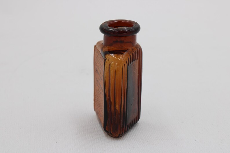 K&ouml;nnte beinhalten: Bernsteinfarbene, rechteckige Glasflasche mit rundem Hals. Die Flasche hat vertikale Rippendetails an den Seiten und eine strukturierte Oberfl&auml;che. Die Flasche ist leer und scheint vintage zu sein.