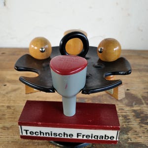 Peut inclure: Un tampon vintage avec une t&ecirc;te rouge et grise, une poign&eacute;e noire et des accents en bois. Le tampon est mont&eacute; sur une base en bois rouge avec le texte "Technische Freigabe" en blanc.