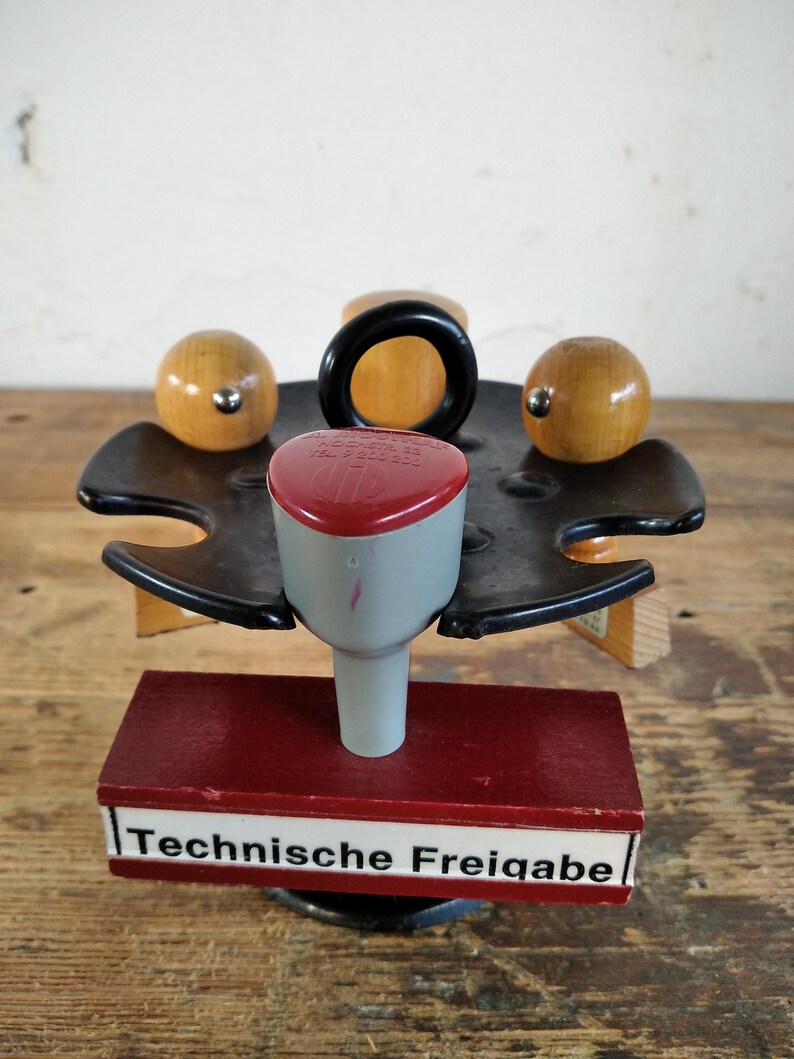 Peut inclure: Un tampon vintage avec une t&ecirc;te rouge et grise, une poign&eacute;e noire et des accents en bois. Le tampon est mont&eacute; sur une base en bois rouge avec le texte "Technische Freigabe" en blanc.