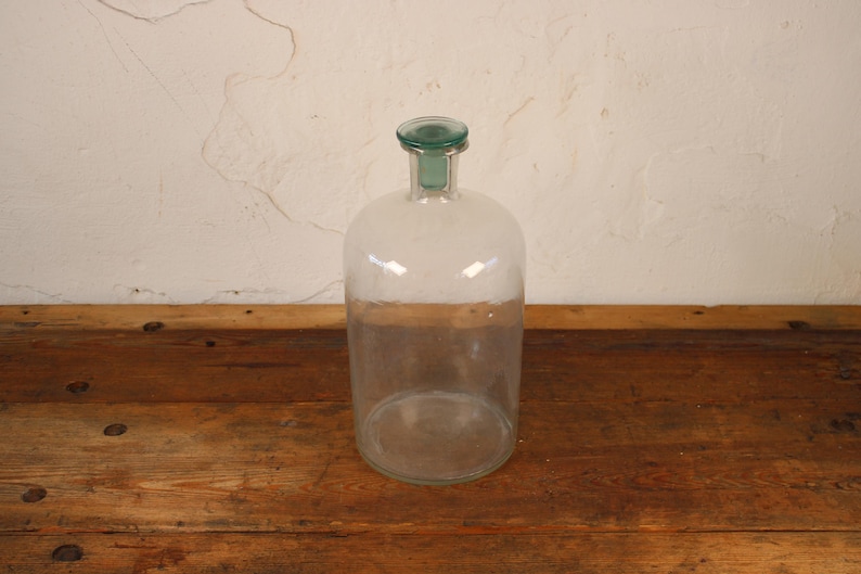 May include: A clear glass bottle with a rounded body and a green glass stopper. The bottle sits on a rustic wooden surface. The background is a textured, off-white wall.