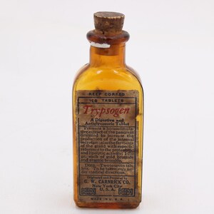 May include: An antique amber glass bottle with a cork stopper. The label reads "Trypsogen" and describes it as a digestive and anti-glycemic tablet. The bottle is marked with "Made in U.S.A."