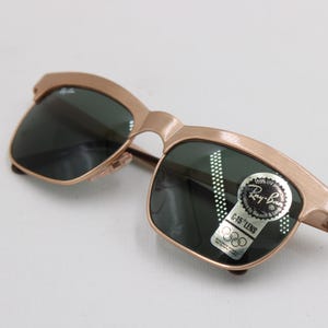 May include: Gold-tone Ray-Ban sunglasses with dark green lenses. The sunglasses have a classic design with a sticker that reads "Ray-Ban G-15 LENS". The frame is a light gold color.