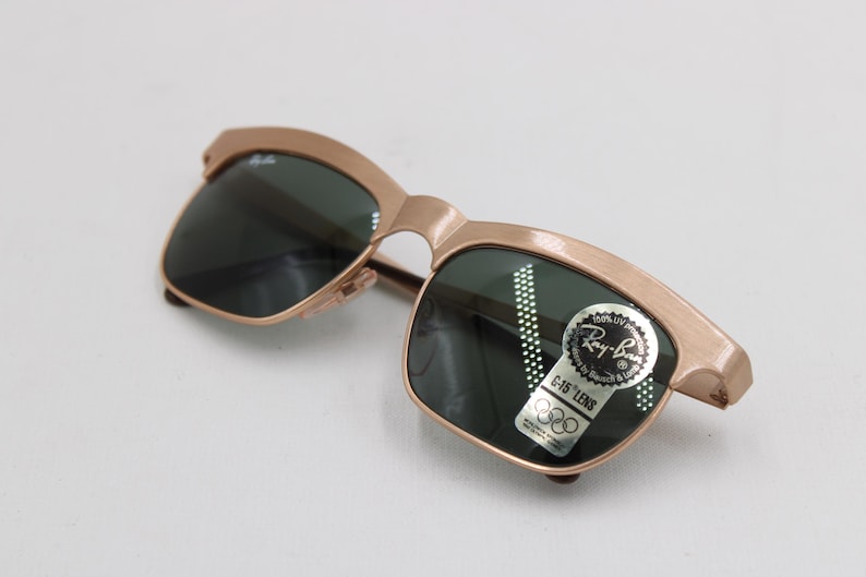 May include: Gold-tone Ray-Ban sunglasses with dark green lenses. The sunglasses have a classic design with a sticker that reads "Ray-Ban G-15 LENS". The frame is a light gold color.