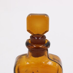 May include: Close-up of a vintage amber glass bottle with a square stopper. The bottle and stopper are a warm, transparent amber color. The stopper has a unique, angular design. The bottle's neck has a dark brown ring.