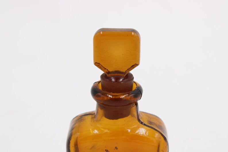 May include: Close-up of a vintage amber glass bottle with a square stopper. The bottle and stopper are a warm, transparent amber color. The stopper has a unique, angular design. The bottle's neck has a dark brown ring.