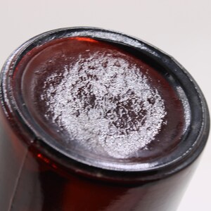 Puede incluir: Primer plano de la base de una botella de vidrio &aacute;mbar oscuro. La base tiene un aspecto texturizado y esmerilado. Los lados de la botella son lisos y cil&iacute;ndricos, con una base ligeramente redondeada.