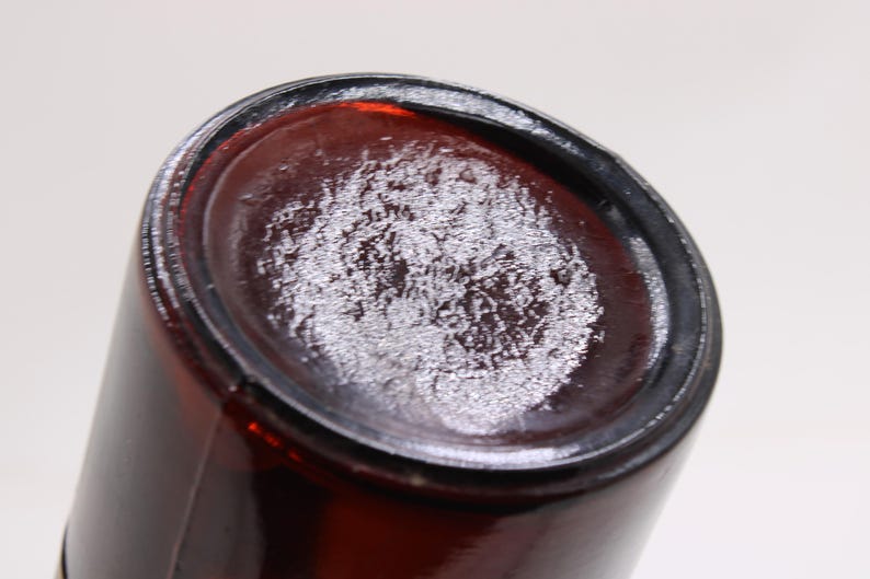 Puede incluir: Primer plano de la base de una botella de vidrio &aacute;mbar oscuro. La base tiene un aspecto texturizado y esmerilado. Los lados de la botella son lisos y cil&iacute;ndricos, con una base ligeramente redondeada.