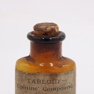 May include: Antique amber glass bottle with a cork stopper. The bottle has a dark brown neck and a label that reads "TABLOID Epinine Compound". The bottle is likely from the early 20th century and is a vintage apothecary item.