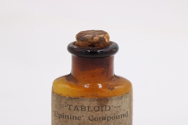 May include: Antique amber glass bottle with a cork stopper. The bottle has a dark brown neck and a label that reads "TABLOID Epinine Compound". The bottle is likely from the early 20th century and is a vintage apothecary item.