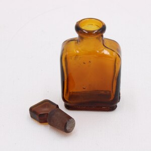 May include: Amber-colored, square-shaped glass bottle with a rounded neck and a separate stopper. The bottle and stopper are made of glass. The bottle is empty and sits on a white surface.