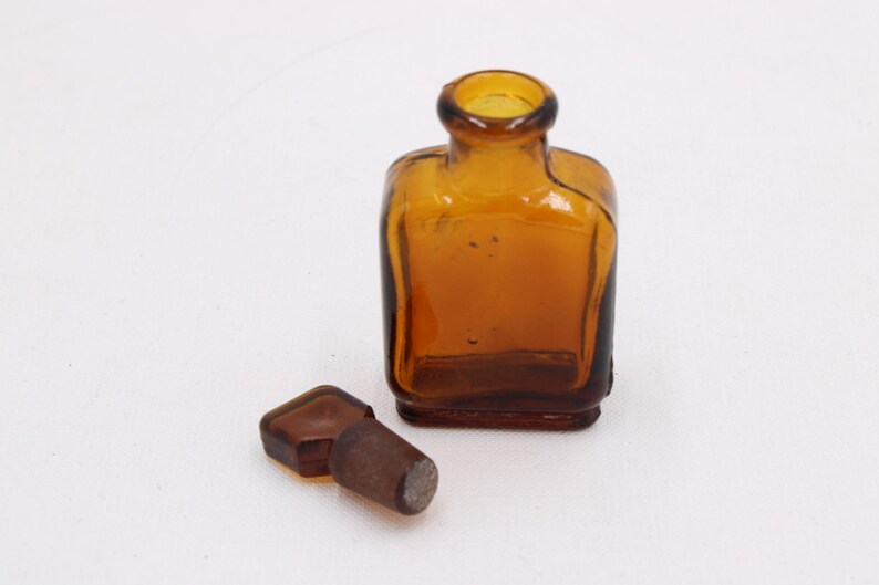 May include: Amber-colored, square-shaped glass bottle with a rounded neck and a separate stopper. The bottle and stopper are made of glass. The bottle is empty and sits on a white surface.
