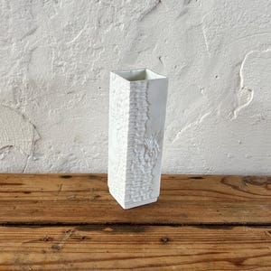 Puede incluir: Un jarrón de cerámica blanco con una superficie texturizada. El jarrón es cuadrado y tiene una abertura rectangular en la parte superior.