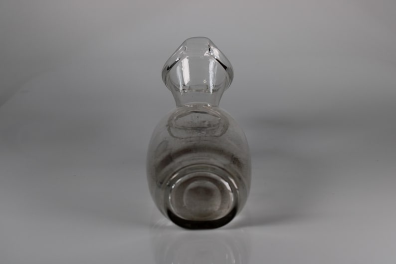 May include: A clear glass decanter with a handle. The decanter has a rounded body and a narrow neck that leads to a handle. The glass is transparent, allowing a view of the contents. The decanter is likely used for serving beverages.
