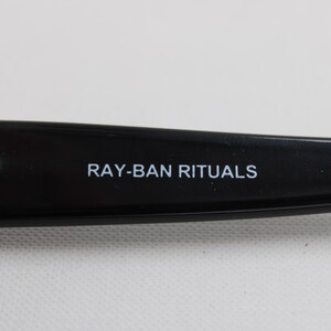 Puede incluir: Primer plano de una patilla de gafas negra con el texto "RAY-BAN RITUALS" en blanco. La bisagra es plateada. Las gafas son probablemente para la correcci&oacute;n de la visi&oacute;n o la protecci&oacute;n solar.