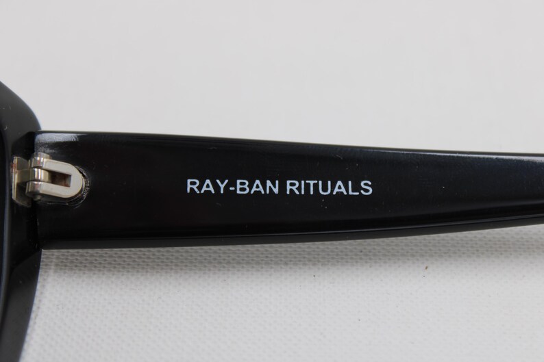 Puede incluir: Primer plano de una patilla de gafas negra con el texto "RAY-BAN RITUALS" en blanco. La bisagra es plateada. Las gafas son probablemente para la correcci&oacute;n de la visi&oacute;n o la protecci&oacute;n solar.