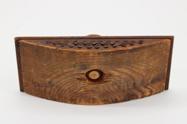 May include: A handcrafted wooden box with a curved front. The box is made of dark brown wood, featuring a carved geometric pattern along the top edge and a circular indentation in the center. The wood grain is visible.