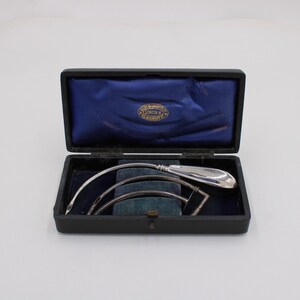 May include: An open black box with a blue satin interior and a silver instrument set. The set includes a curved, elongated tool with a rounded handle, and several other curved metal pieces. The box is on a white surface.
