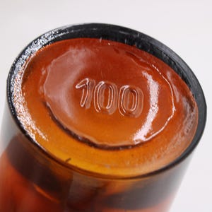 May include: Close-up of the bottom of a brown glass bottle. The number "100" is embossed on the base. The bottle has a dark rim and a glossy, amber-colored interior.