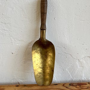 Puede incluir: Una pala de latón vintage con mango de madera. La pala es de color dorado claro y tiene una forma ligeramente redondeada. El mango es de color marrón oscuro y tiene un diseño sencillo.