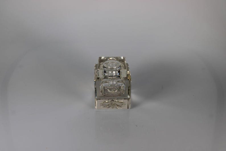 May include: A rectangular, clear glass inkwell with a silver-colored metal frame. The inkwell has a hinged lid and a decorative floral design on the front. The inkwell is likely vintage and suitable for calligraphy.