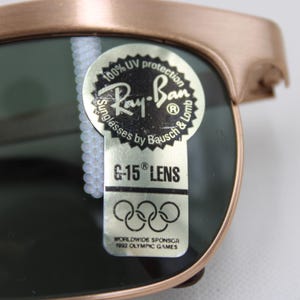 May include: Close-up of Ray-Ban sunglasses with a bronze frame and dark green lenses. The sunglasses have a sticker that reads "100% UV protection" and "G-15 LENS". The sticker also includes the Ray-Ban logo and text about the 1992 Olympic Games.