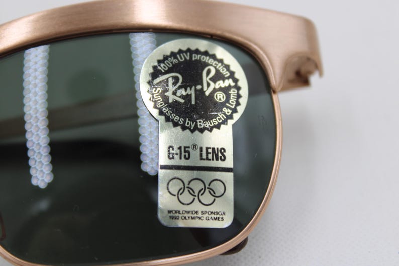 May include: Close-up of Ray-Ban sunglasses with a bronze frame and dark green lenses. The sunglasses have a sticker that reads "100% UV protection" and "G-15 LENS". The sticker also includes the Ray-Ban logo and text about the 1992 Olympic Games.