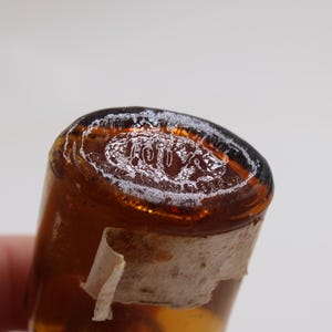 Puede incluir: Primer plano de una botella de vidrio &aacute;mbar vintage con la parte superior redondeada. La parte superior tiene una inscripci&oacute;n blanca descolorida. Un trozo de una etiqueta rasgada y envejecida est&aacute; adherido a la botella.