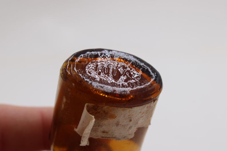 Puede incluir: Primer plano de una botella de vidrio &aacute;mbar vintage con la parte superior redondeada. La parte superior tiene una inscripci&oacute;n blanca descolorida. Un trozo de una etiqueta rasgada y envejecida est&aacute; adherido a la botella.
