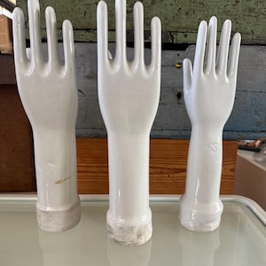 Könnte beinhalten: Drei weiße Keramikhandformen, jede mit einer detaillierten Darstellung von Fingern und einem Handgelenkansatz. Die Hände sind in einer Reihe angeordnet und zeigen ihre glatte, glänzende Oberfläche. Der Hintergrund zeigt eine Mischung aus Holzpaneelen und einer reflektierenden Oberfläche.